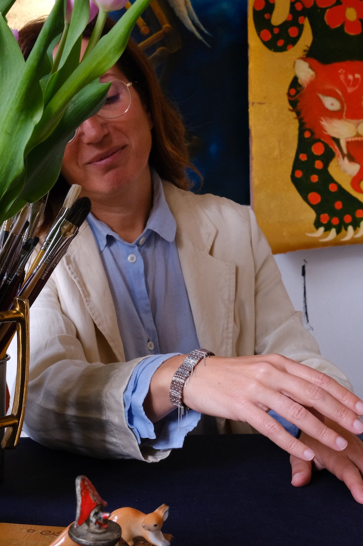 Lifestyle shot of a mocha-colored minimalist woven bracelet on a wrist, surrounded by artistic studio elements like paintbrushes and floral accents.